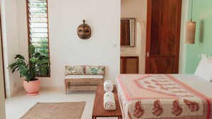Una habitación con cama, un banco, una planta en maceta y un máscara colgada en la pared.