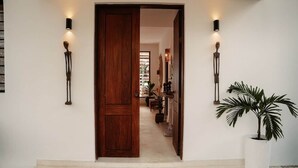 Un couloir avec une porte en bois, deux appliques murales et une plante en pot.