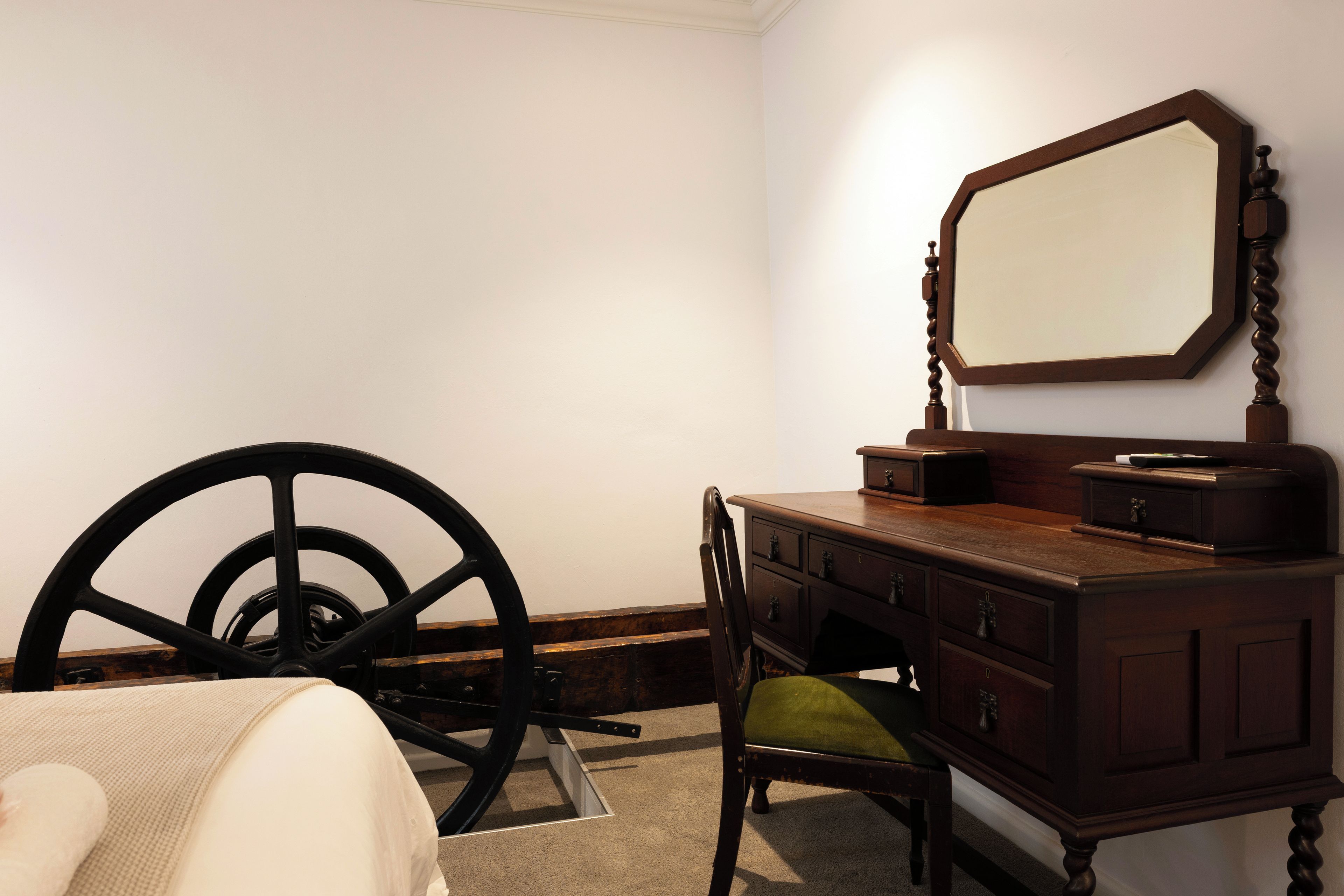 A room with a wooden dresser, a chair, a large mirror, and a large wheel.