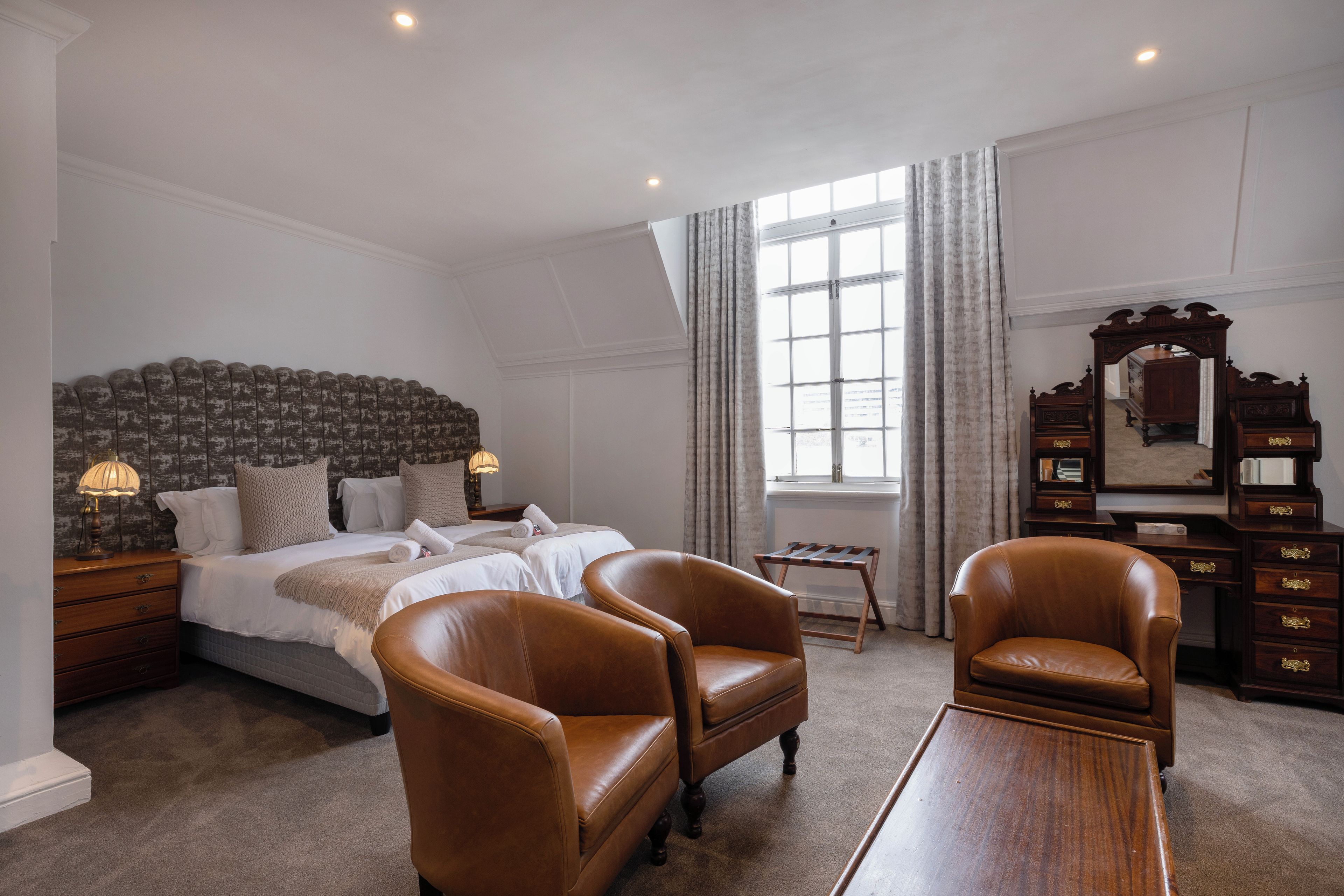 Una habitación de hotel con una cama grande, dos sillones de cuero, un tocador de madera y un espejo.