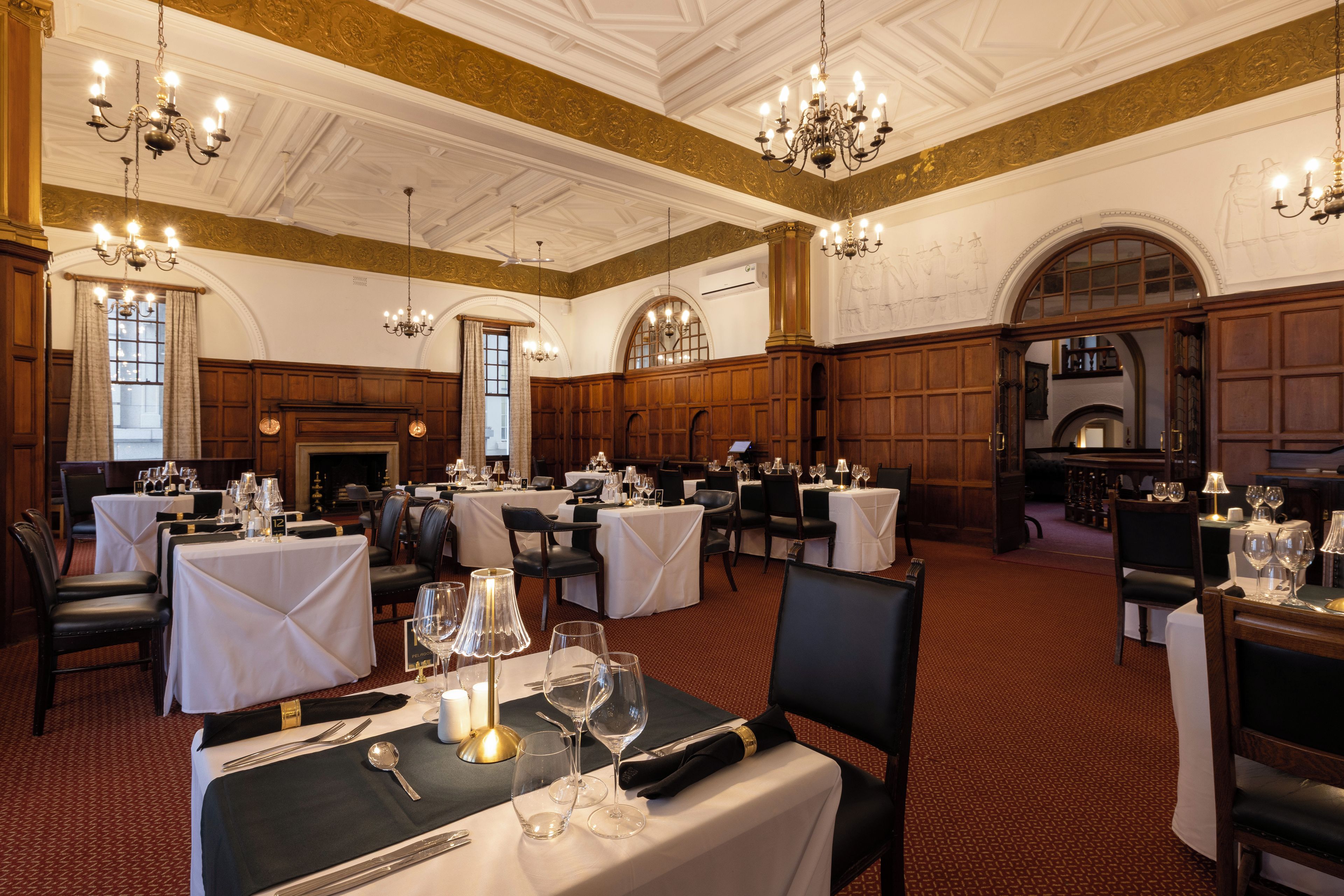 Une salle à manger élégante, avec plusieurs tables dressées pour le service, des lustres et un plafond haut orné de moulures décoratives.
