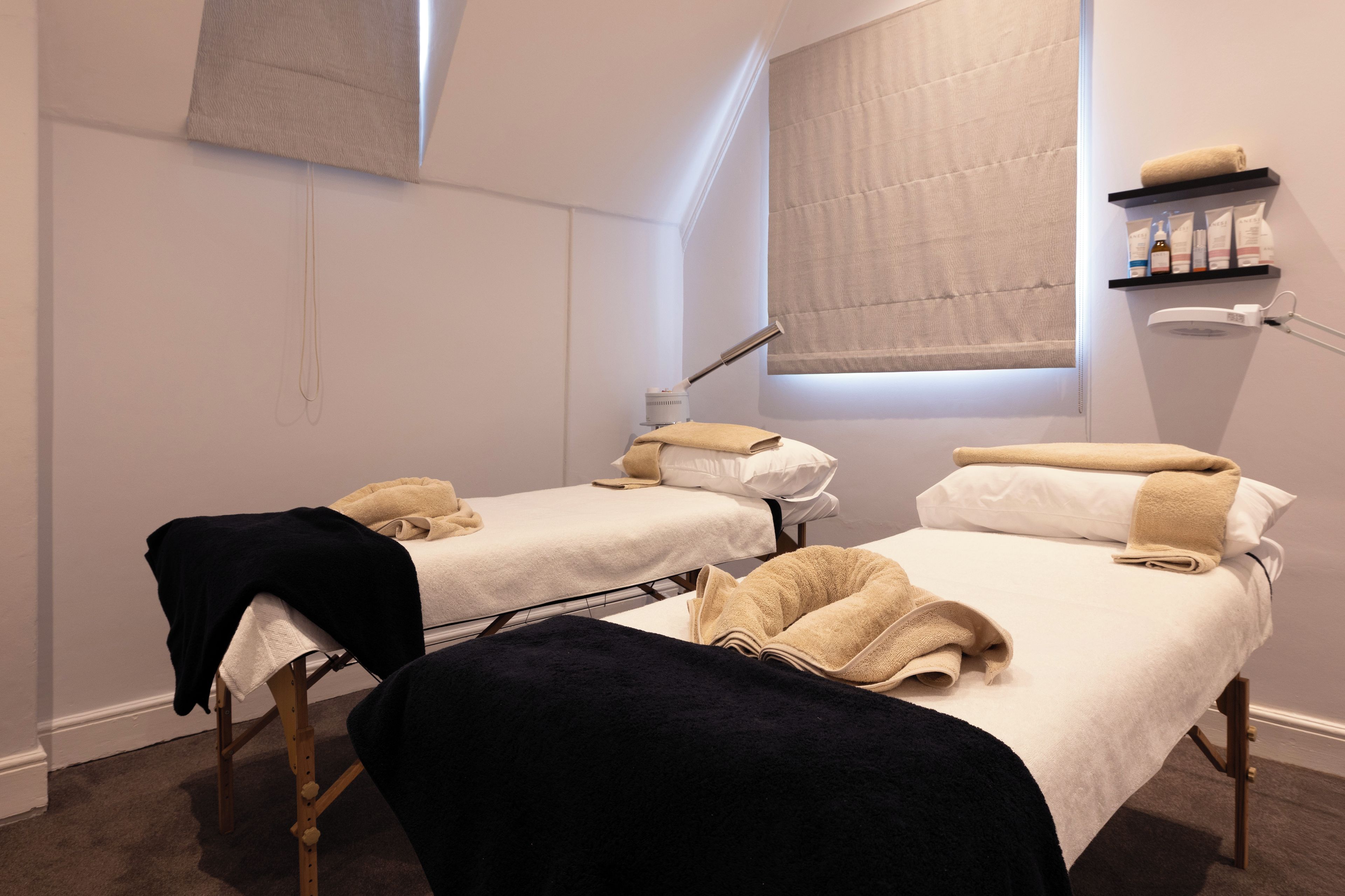 A room with two treatment beds, a window with a blind, a shelf with products, and a desk lamp.