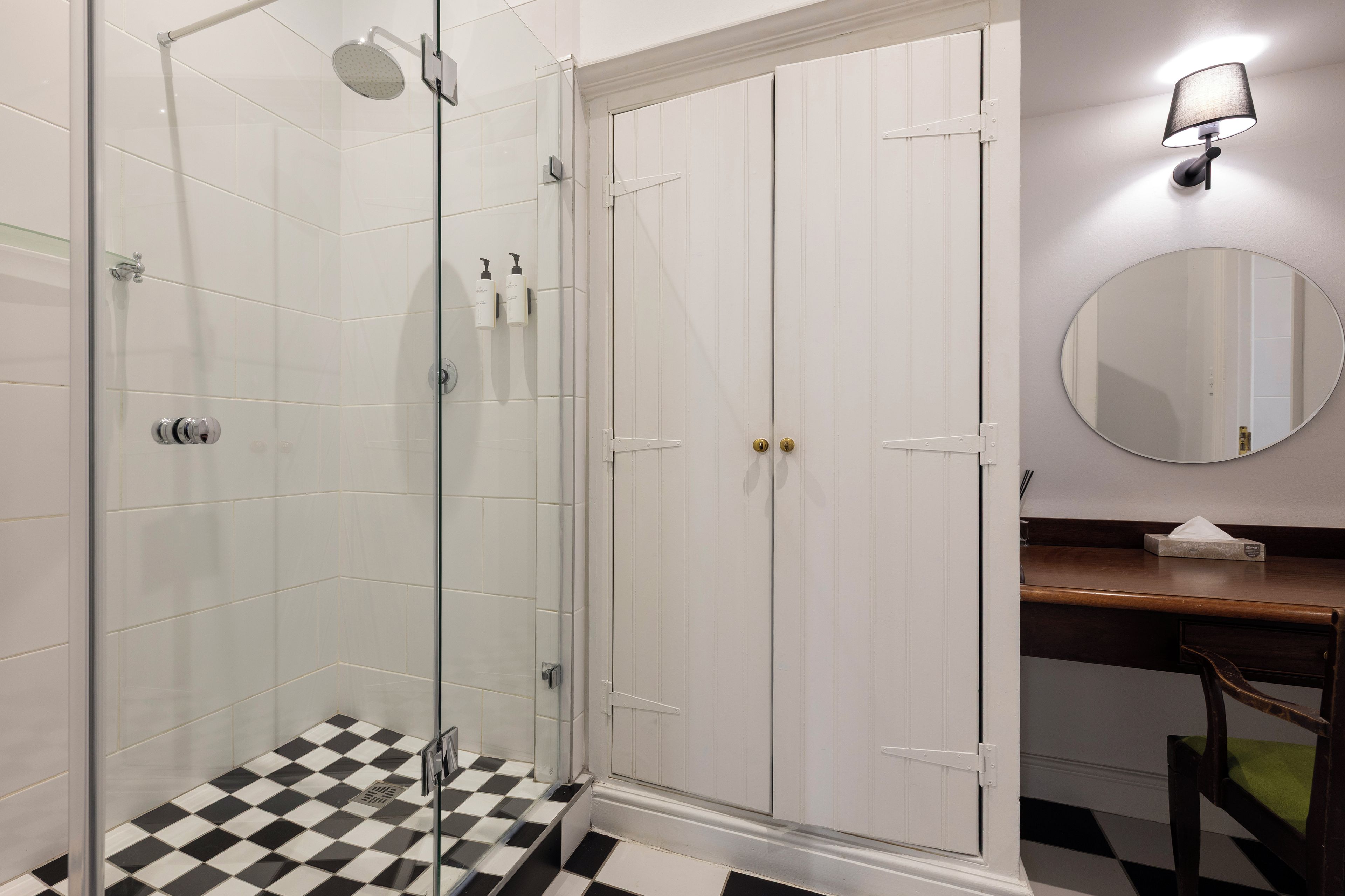 A bathroom with a glass shower enclosure, a white tiled floor, a wooden vanity with a mirror, and a white door with a gold handle.