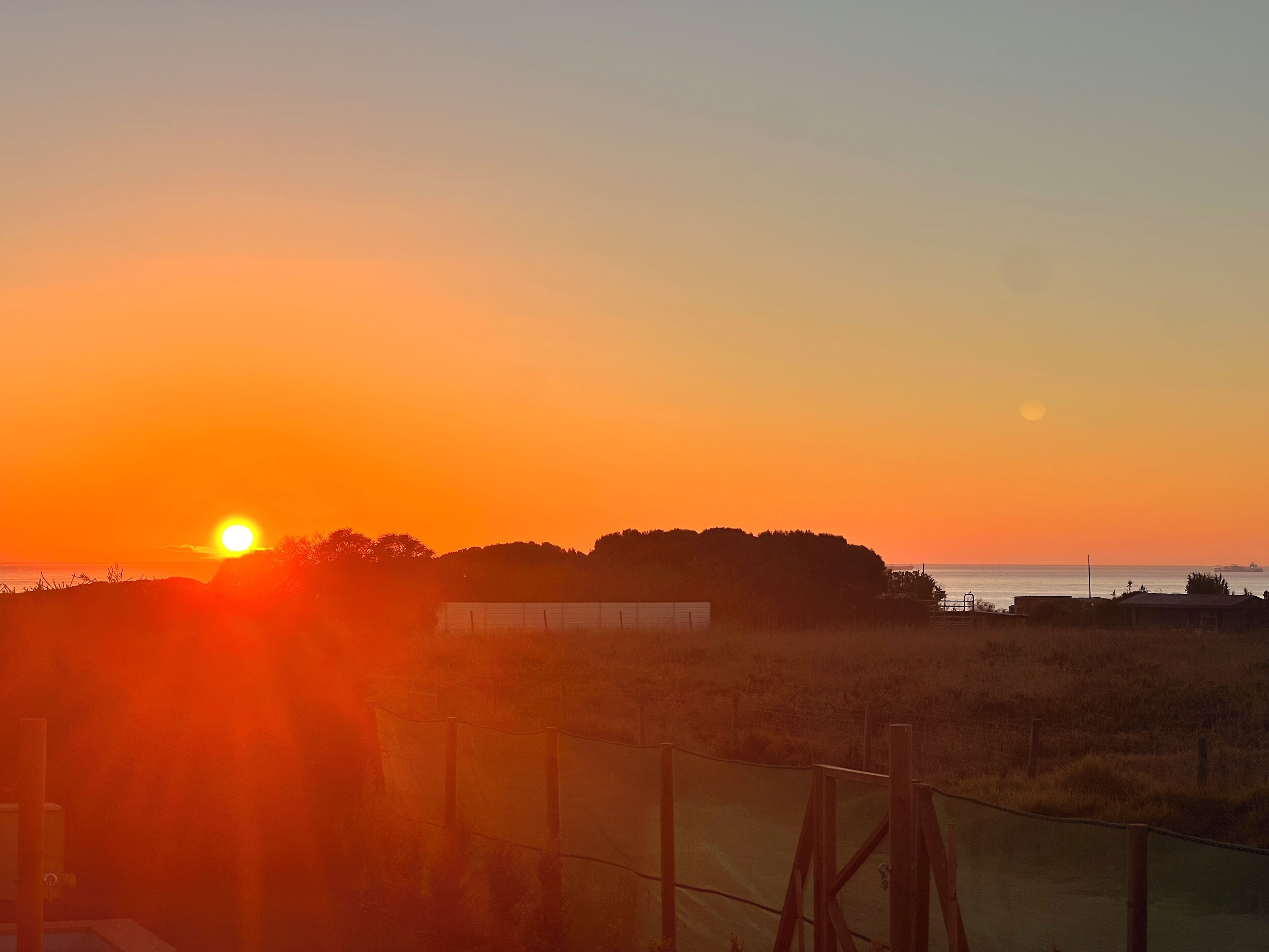 A sunset over a coastal landscape with a fence and vegetation.
