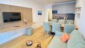 A modern living room with a flat-screen TV, a dining area with light blue chairs, and a kitchen in the background.