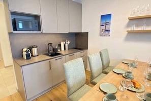 Cocina compacta con microondas, hervidor y utensilios, una mesa de comedor con sillas, y una estantería de pared con vasos.