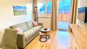 Un salón con sofá, una mesita redonda, un banco de madera, un televisor montado en la pared y un balcón con vistas a las montañas.