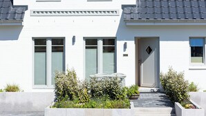 Un edificio blanco de dos pisos con techo gris, una puerta principal y un pequeño jardín con plantas y un banco.