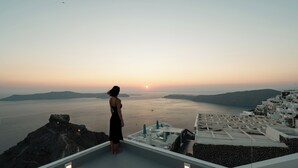 Eine Frau steht auf einem Dach und blickt auf eine Küstenlandschaft bei Sonnenuntergang.