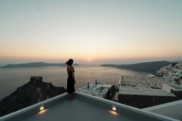 A woman standing on a rooftop overlooking a coastal landscape with a sunset.