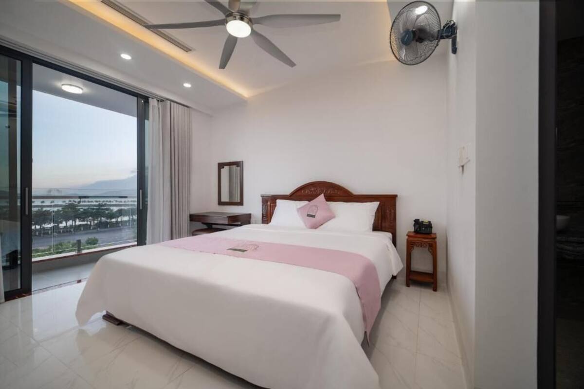 Ein Hotelzimmer mit einem großen Bett, einem Deckenventilator, einem Wandventilator, einem Nachttisch und Blick auf eine Stadtlandschaft durch große Fenster.