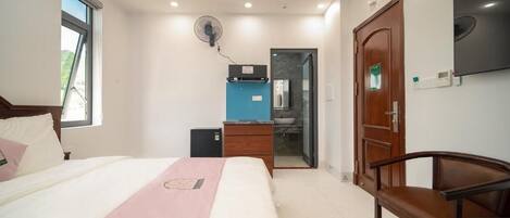 Un dormitorio con cama, escritorio, silla, armario, televisor y baño visible a través de una puerta abierta.