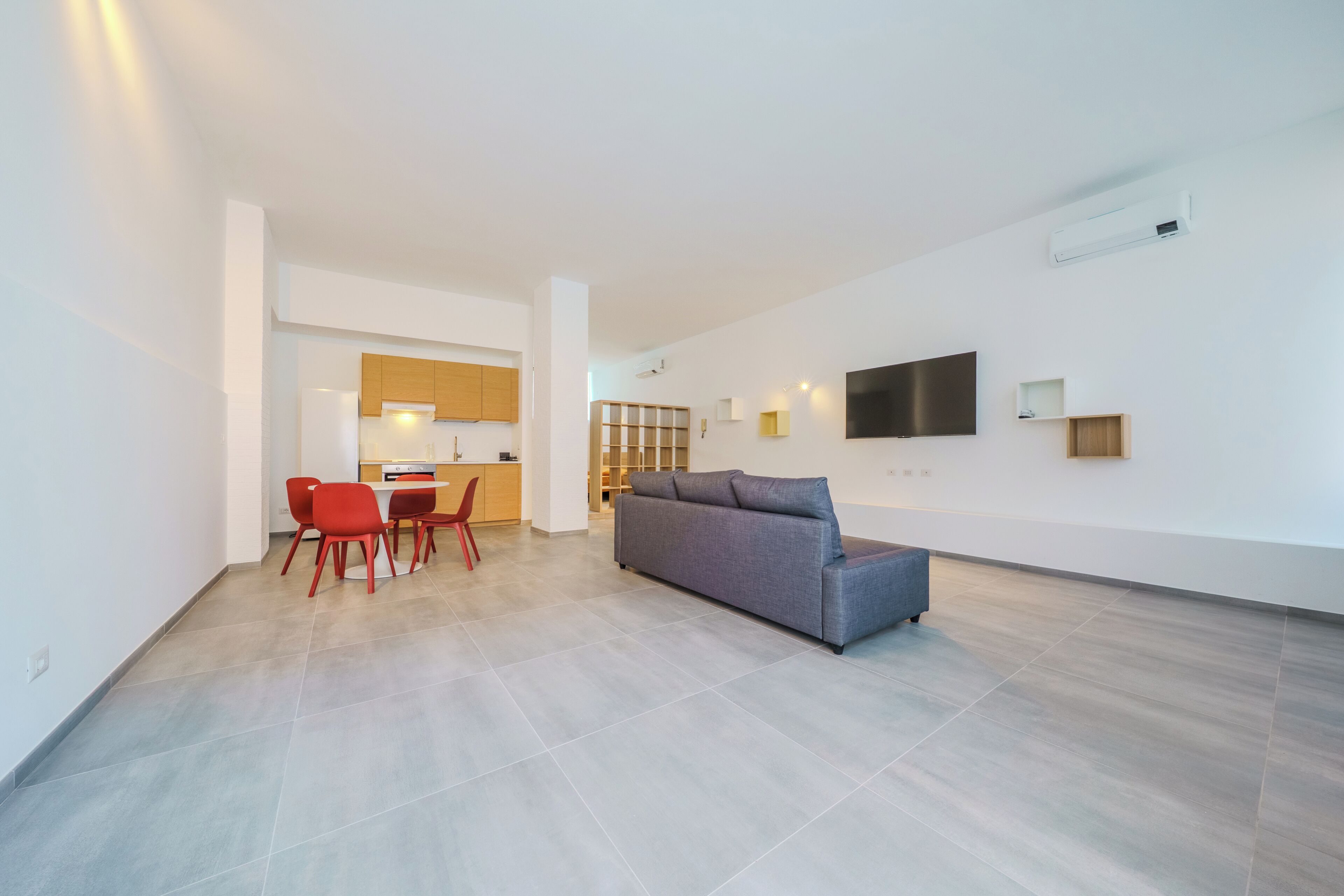 A modern living room with a grey sofa, red chairs, a flat-screen TV, and a kitchen in the background.