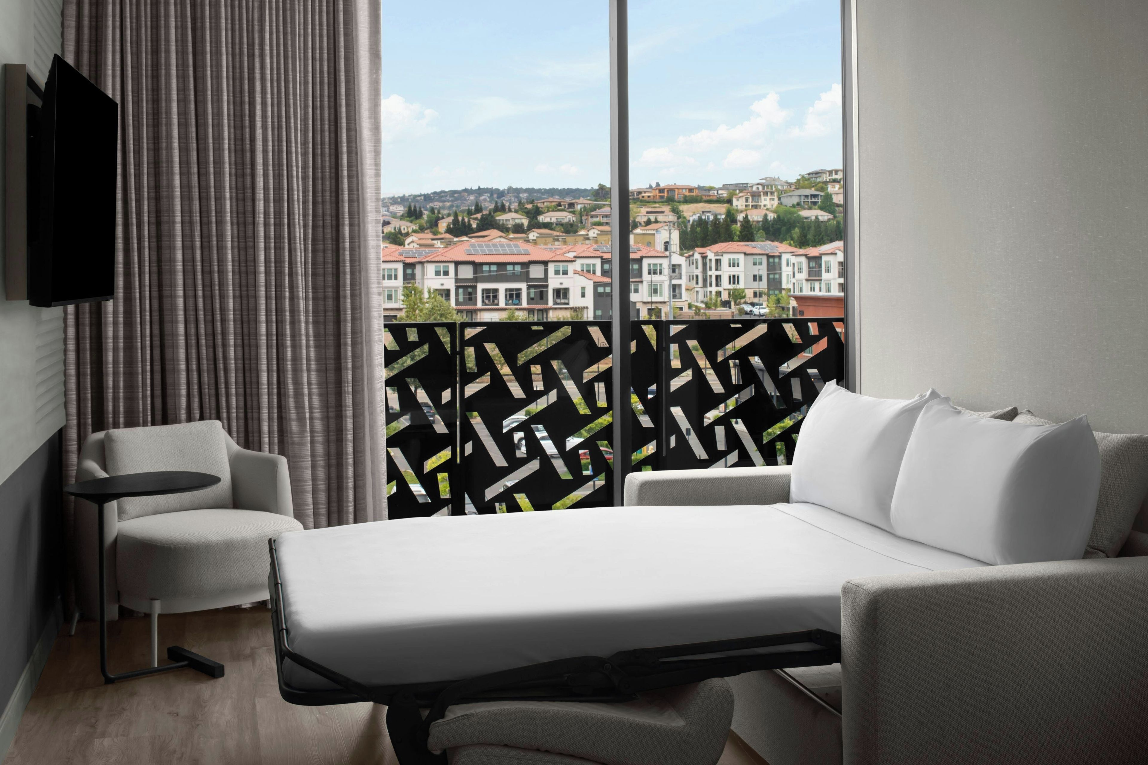 A hotel room with a bed, a sofa, a TV, and a view of a residential area.