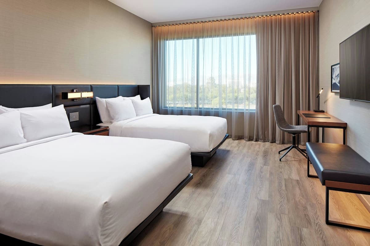 A hotel room with two beds, a desk, a chair, a TV, and a large window with curtains.