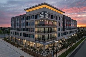 Un hotel de varios pisos con un llamativo letrero de AC AC Hotels Marriott, mesas al aire libre y un estacionamiento.
