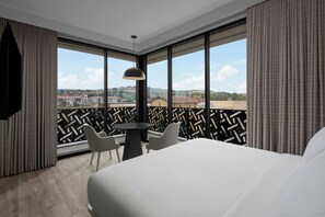 Ett hotellrum med en stor säng, ett runt bord med två stolar, en hängande lampa och utsikt över en stadspanorama genom stora fönster.