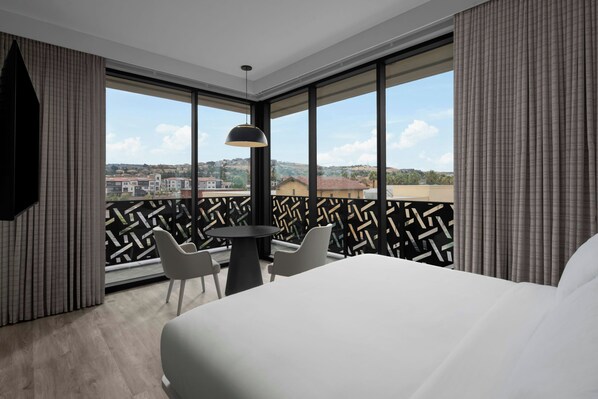 Habitación de hotel con una cama grande, una mesa redonda con dos sillas, una lámpara colgante y vistas a un paisaje urbano a través de amplias ventanas.