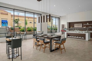 A modern dining area with a large table and chairs, a kitchen area with a buffet, and a view of a building with a blue awning. - AC Hotels by Marriott Sacramento Folsom (Folsom)