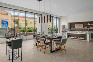 Un comedor moderno con una gran mesa y sillas, una zona de cocina con bufé, y vista a un edificio con toldo azul.