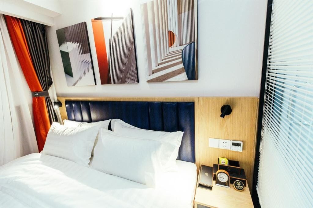 A hotel room with a bed, artwork on the wall, a clock, and a control panel.