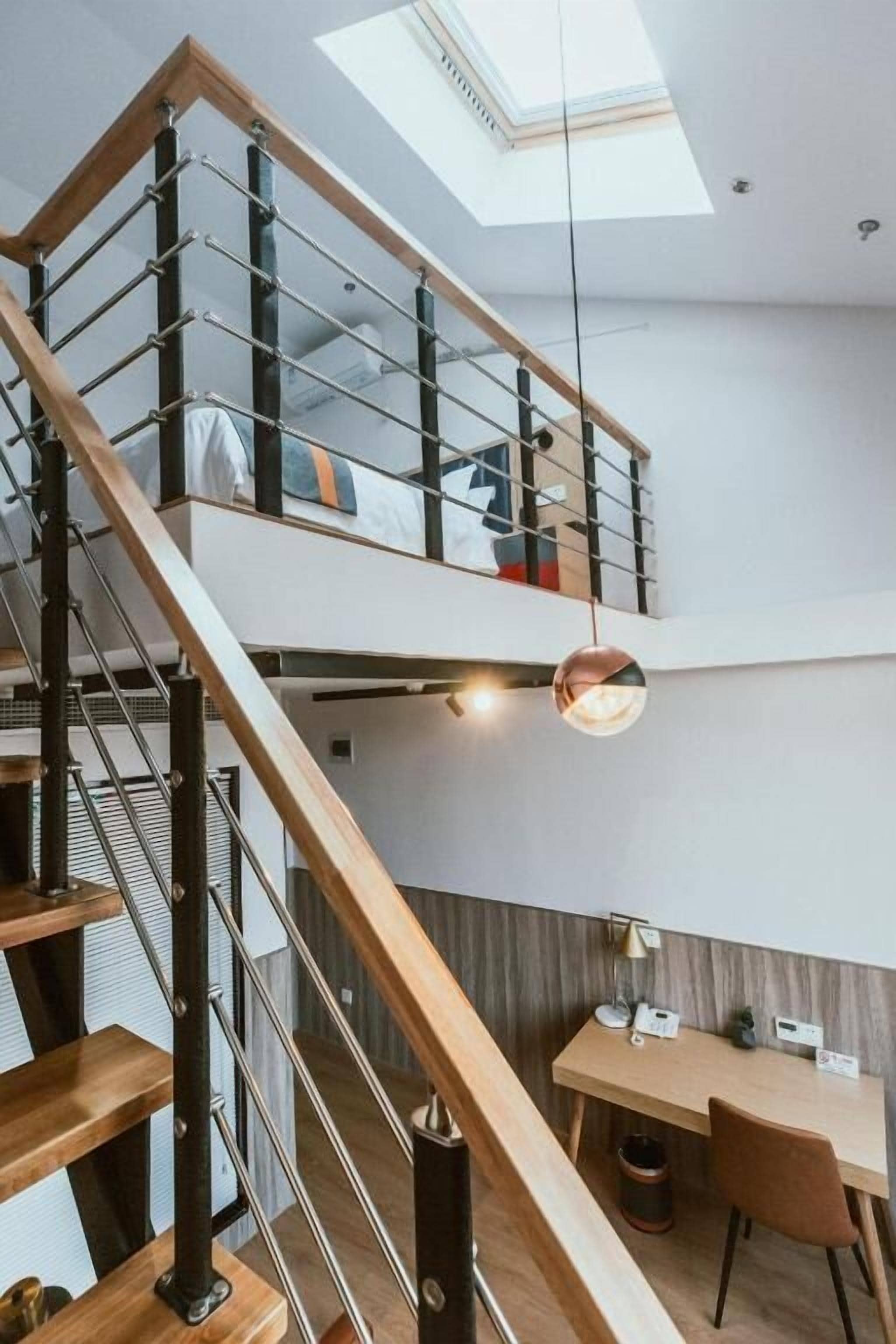 Un intérieur moderne avec un escalier en bois, une mezzanine comprenant un lit, un bureau avec une chaise et un luminaire suspendu.