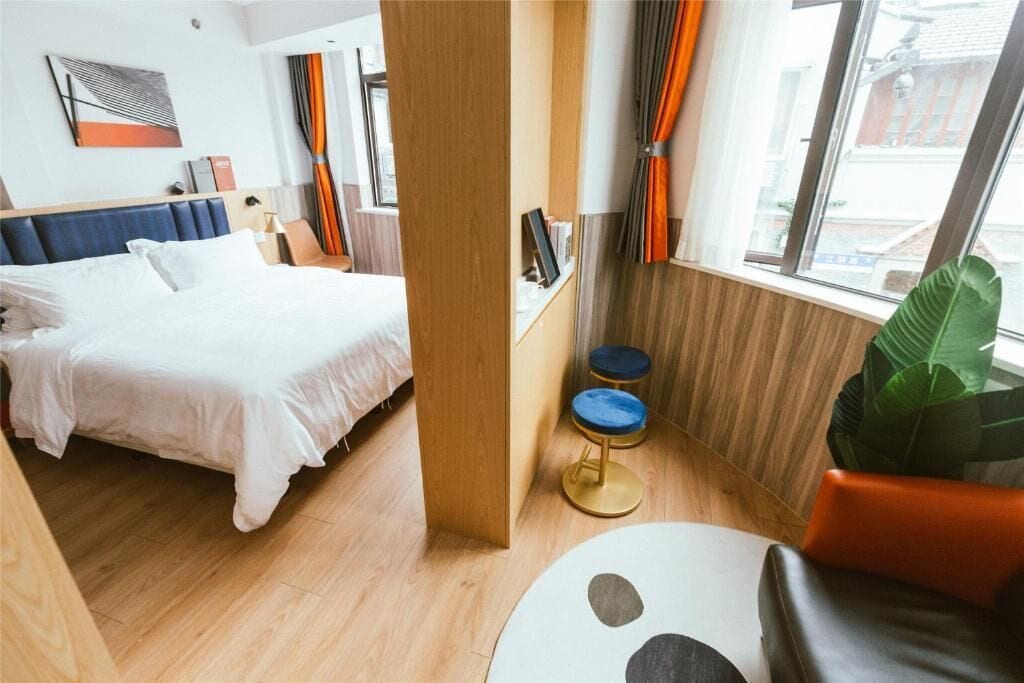 A hotel room with a bed, a desk, a chair, a large window, and a plant.