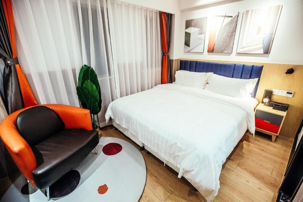 Quarto de hotel com cama, cadeira, mesa de cabeceira, abajur e janela com cortinas.
