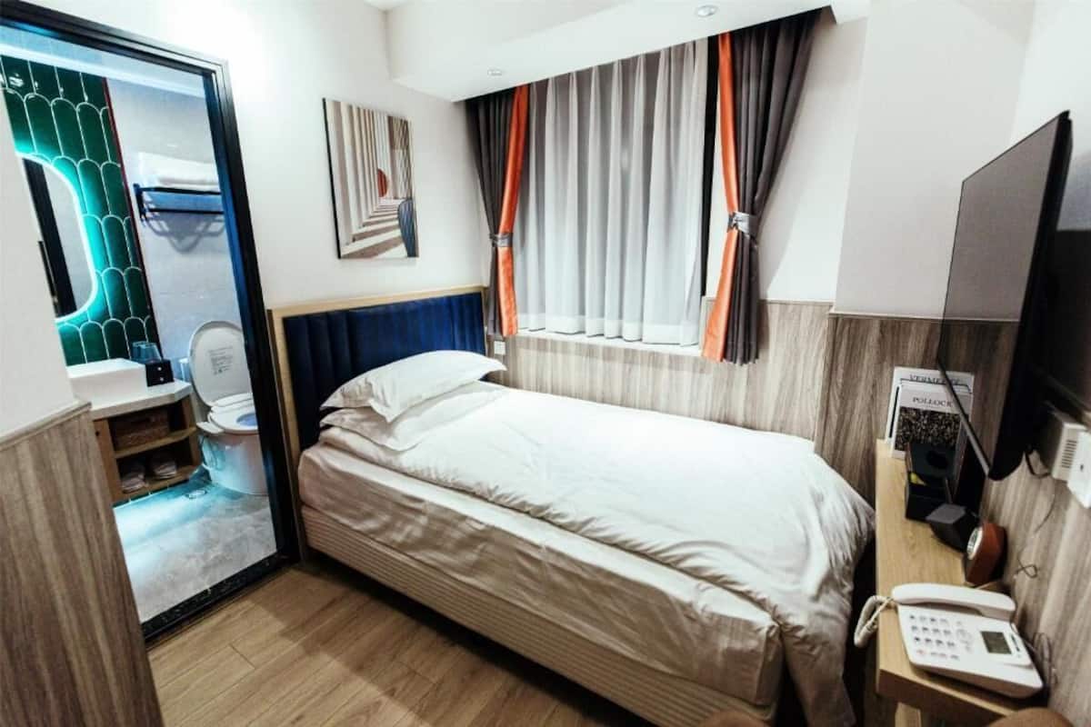 Een kleine hotelkamer met een bed, een bedtafeltelefoon, een raam met gordijnen, een spiegel en een kleine badkamer die door een geopende deur zichtbaar is.