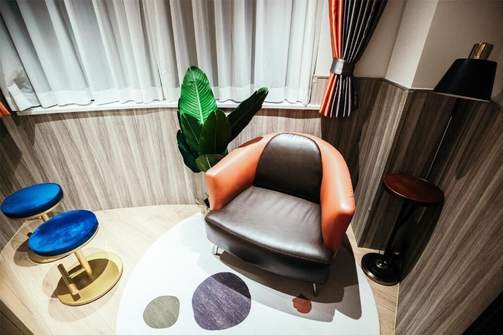 A modern room with a leather armchair, two blue bar stools, a potted plant, and a window with curtains.