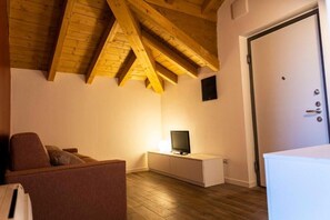 A room with a sloped ceiling, a television on a white cabinet, a door with a handle, and a brown couch.