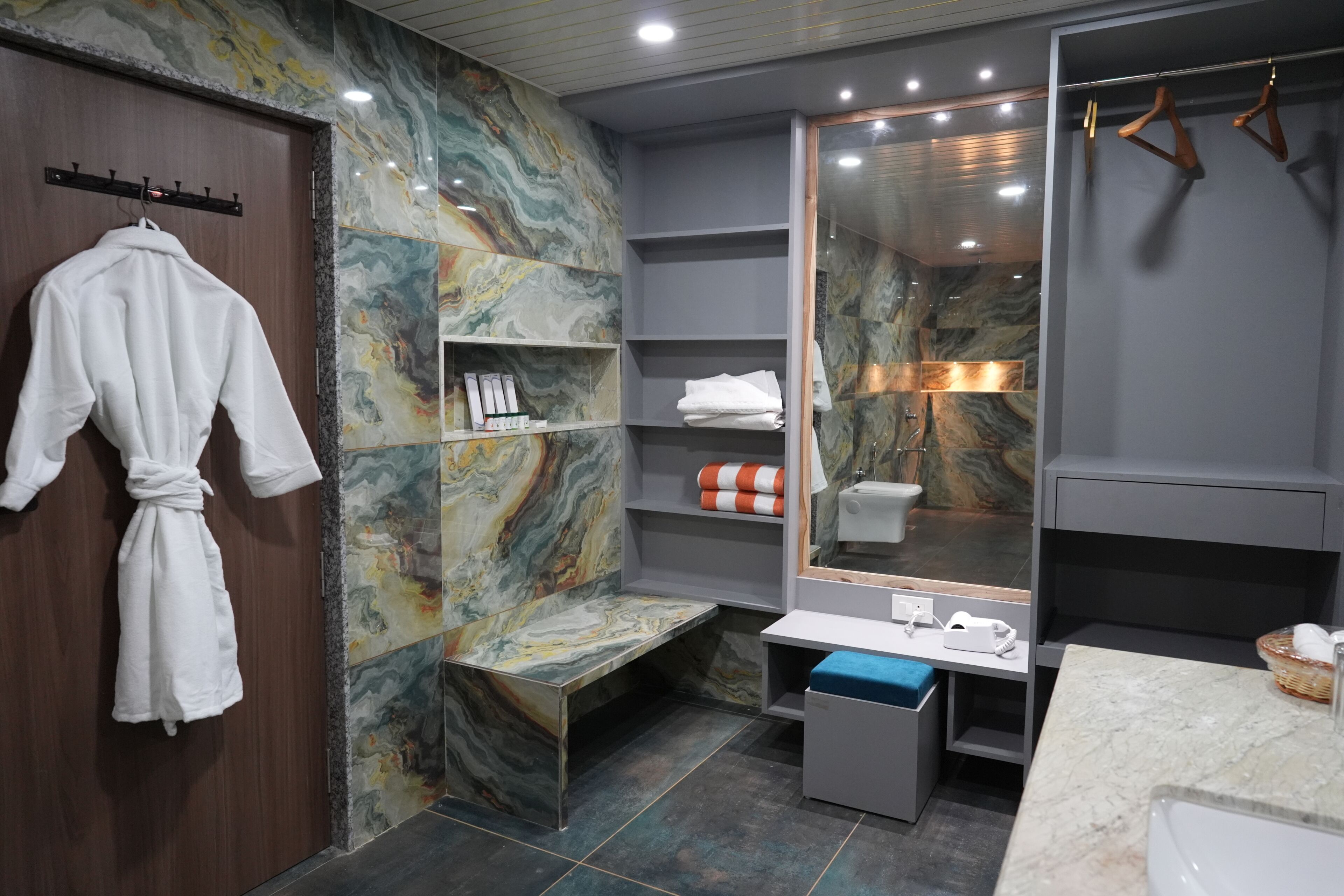 A modern bathroom with a marble wall, a bench, a vanity with a mirror, and a robe rack.