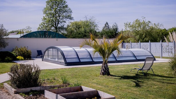 Una estructura de invernadero con techo curvo, una pequeña palmera y una silla en un jardín.