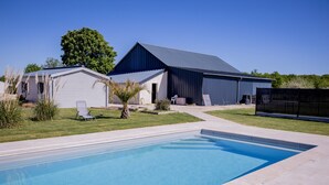 Una piscina con superficie azul cristalina, una silla de descanso blanca y una estructura grande tipo granero con techo oscuro.