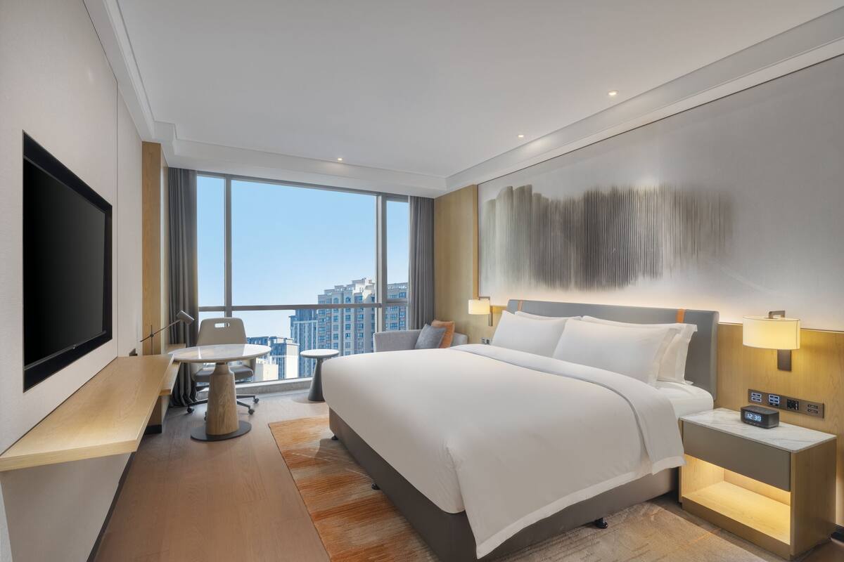 A modern hotel room with a large bed, a desk, a TV, and a city view.