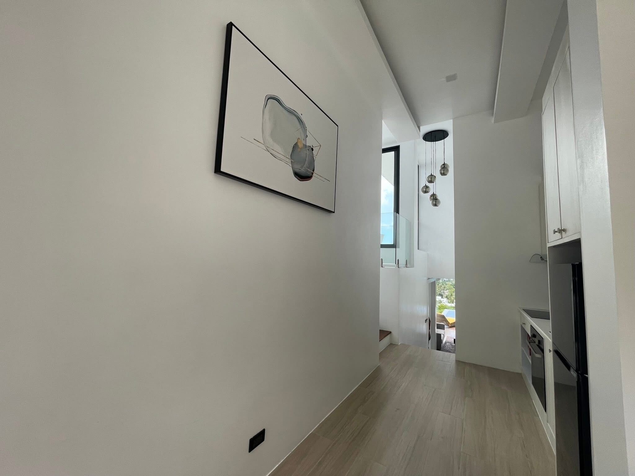 A modern interior with a hallway leading to a kitchen area, featuring a framed artwork, pendant lighting, and a visible window.