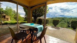 A covered patio with a table and chairs, large windows, and a view of the landscape. - Villa Dorlot (Cazals)