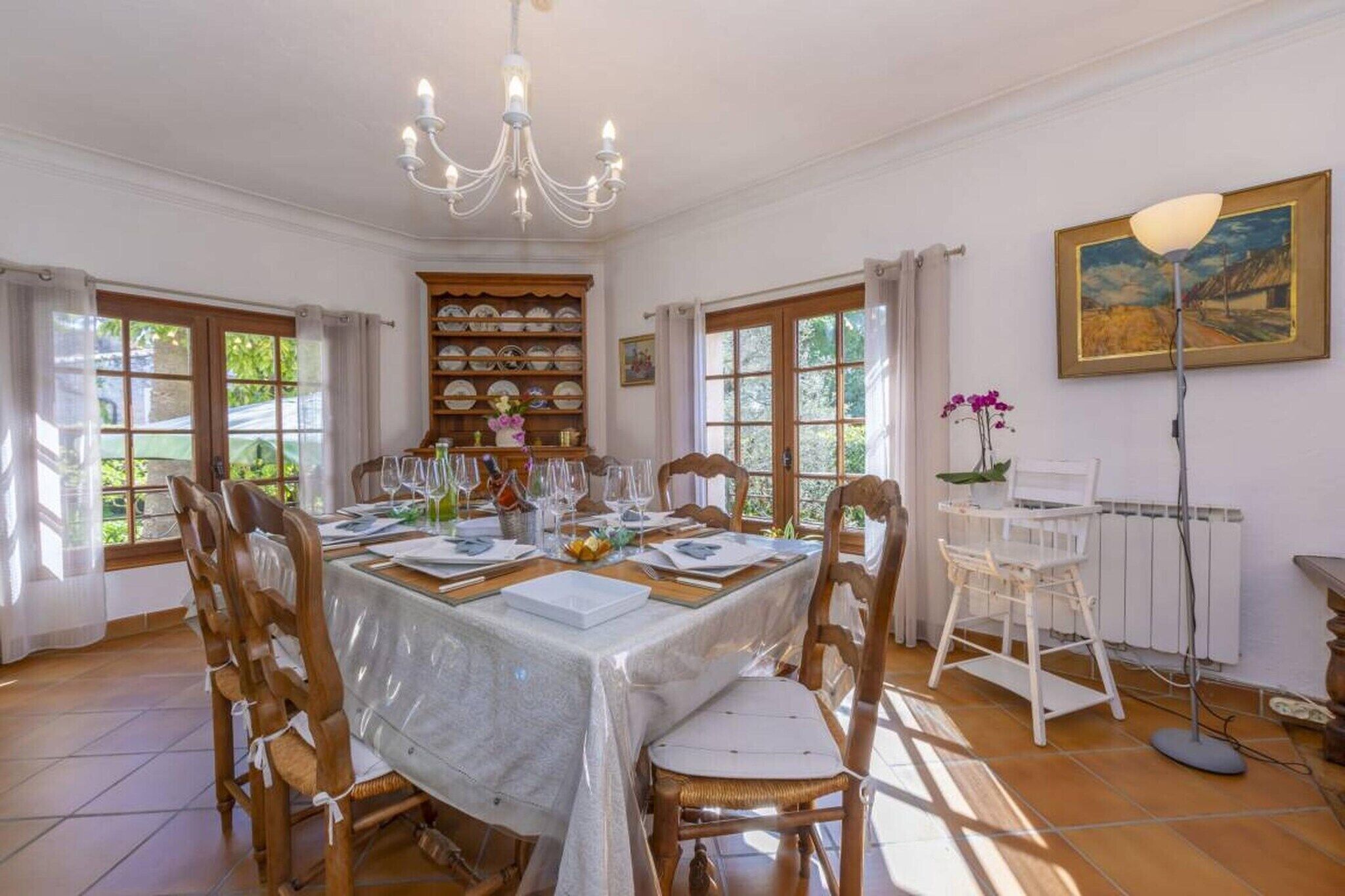 Une salle à manger avec une table dressée pour six personnes, des chaises en bois, un lustre et une vue sur la verdure à l’extérieur.