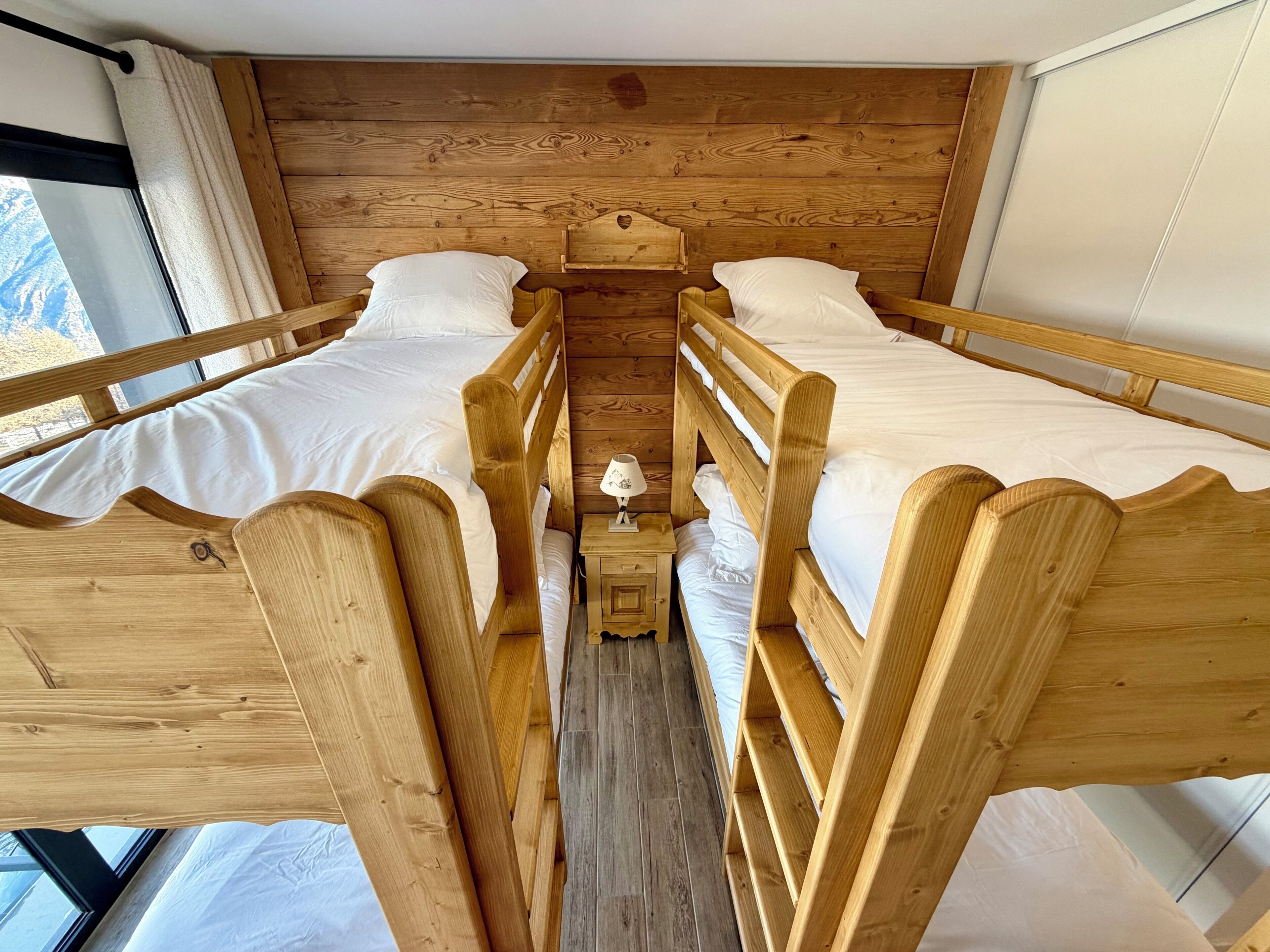 A room with two wooden bunk beds, a small bedside table with a lamp, and a window with curtains.