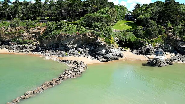 Eine Küstenlandschaft mit einem felsigen Strand, einer kleinen, auf Stelzen gebauten Struktur und einem Gebäude in der Ferne.