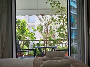Ein Zimmer mit einem Bett, einem kleinen Tisch und einem Stuhl. Es gibt einen Balkon mit Pflanzen und Blick auf Gebäude.