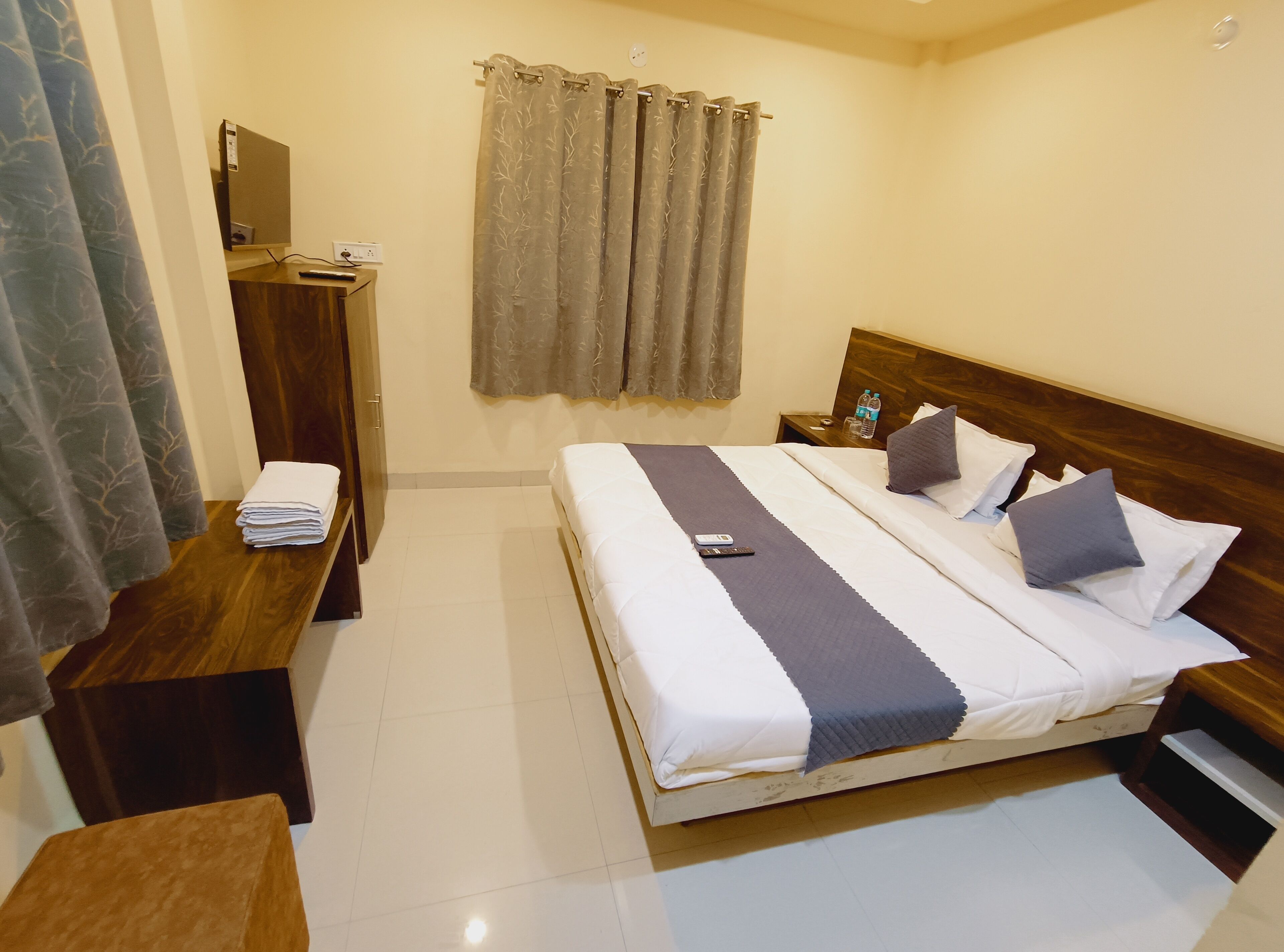 A hotel room with a bed, bedside table, a desk with a chair, a mirror, and a window with curtains.