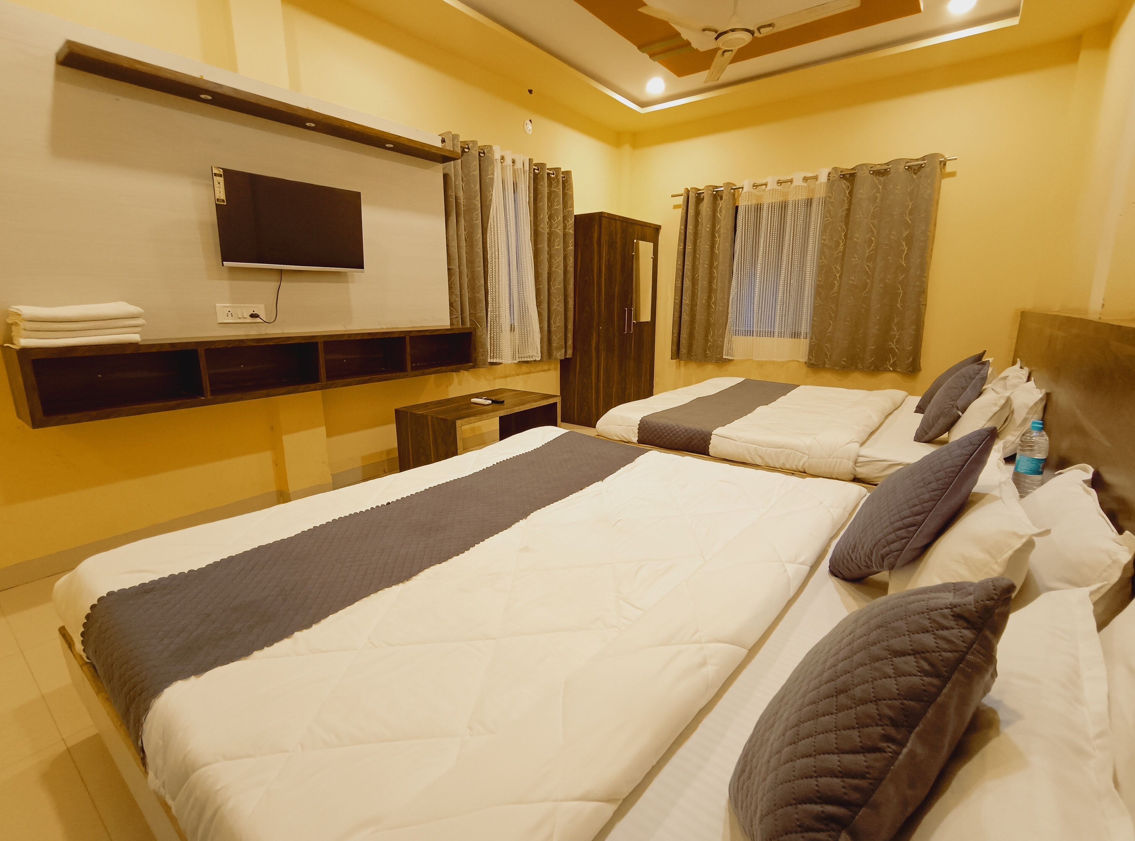 Quarto de hotel com duas camas, televisão de tela plana, mesa de cabeceira de madeira, guarda-roupa e ventilador de teto.