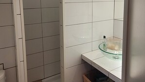 A bathroom with white tiled walls, a glass bowl sink, a wicker basket, and a shower area. - T2 cabin apartment on garden level with pedestrian access to the beach (Agde)
