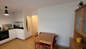 A compact kitchen with white cabinets, a black oven, a wooden table, and red chairs. - T2 cabin apartment on garden level with pedestrian access to the beach (Agde)
