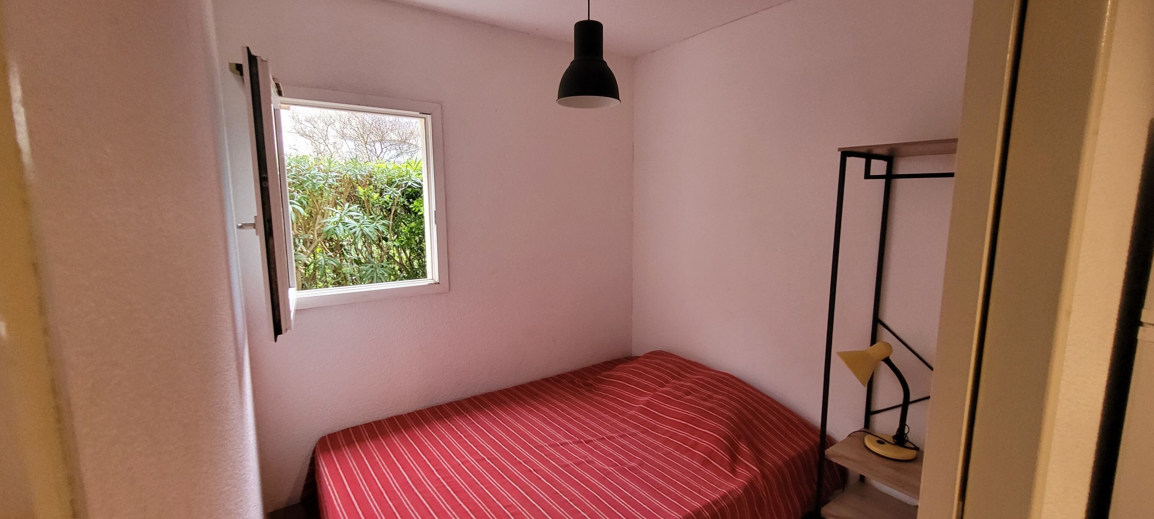 A small, single-bed room with a window, a hanging light fixture, and a small table with a chair.