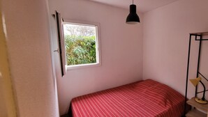 A small, single-bed room with a window, a hanging light fixture, and a small table with a chair. - T2 cabin apartment on garden level with pedestrian access to the beach (Agde)