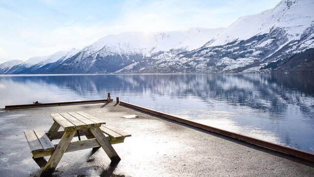 A tranquil lakeside setting with a wooden picnic table and a dock extending into a calm lake, surrounded by snow-capped mountains.