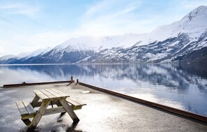 A tranquil lakeside setting with a wooden picnic table and a dock extending into a calm lake, surrounded by snow-capped mountains. - Beach front home in Hovland with sauna (Hovland)