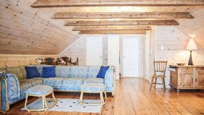 A cozy attic room with a wooden ceiling, a patterned sofa, two matching ottomans, a wooden chair, and a cabinet.